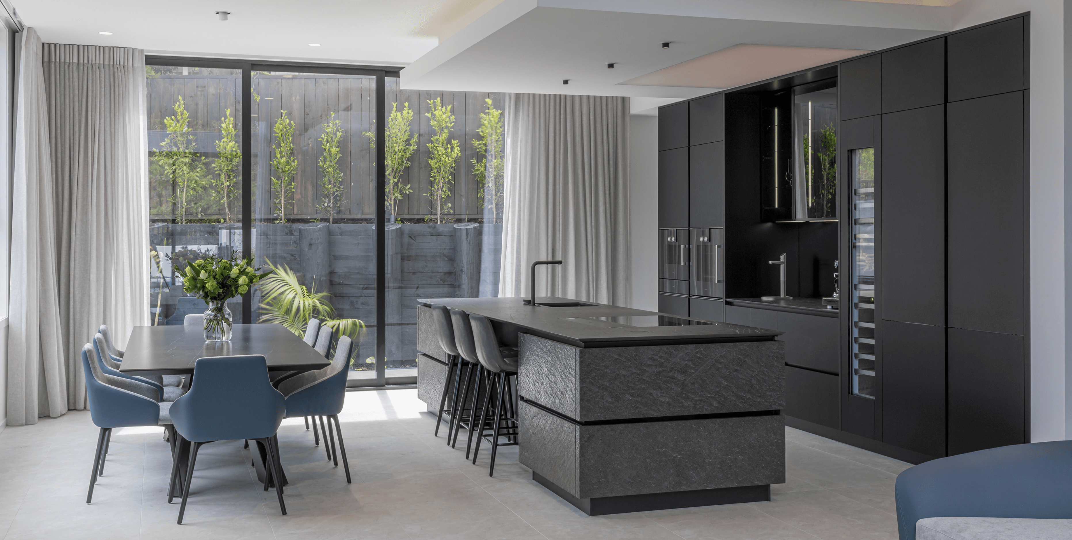 Monochrome kitchen renovation combining matte black finishes, tactile stone and seamless dining integration.