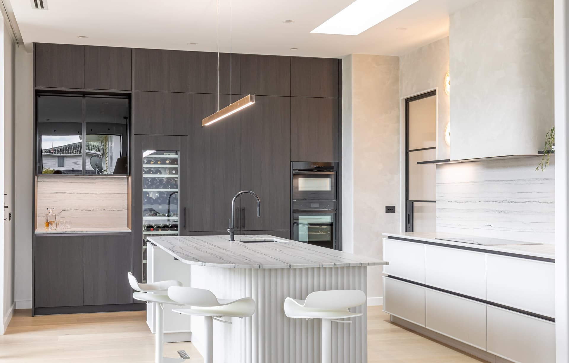 Fluted island and stools in front of the full dark cabinet wall, white cooktop run to the right, in a Westmere home | Kitchens New Zealand