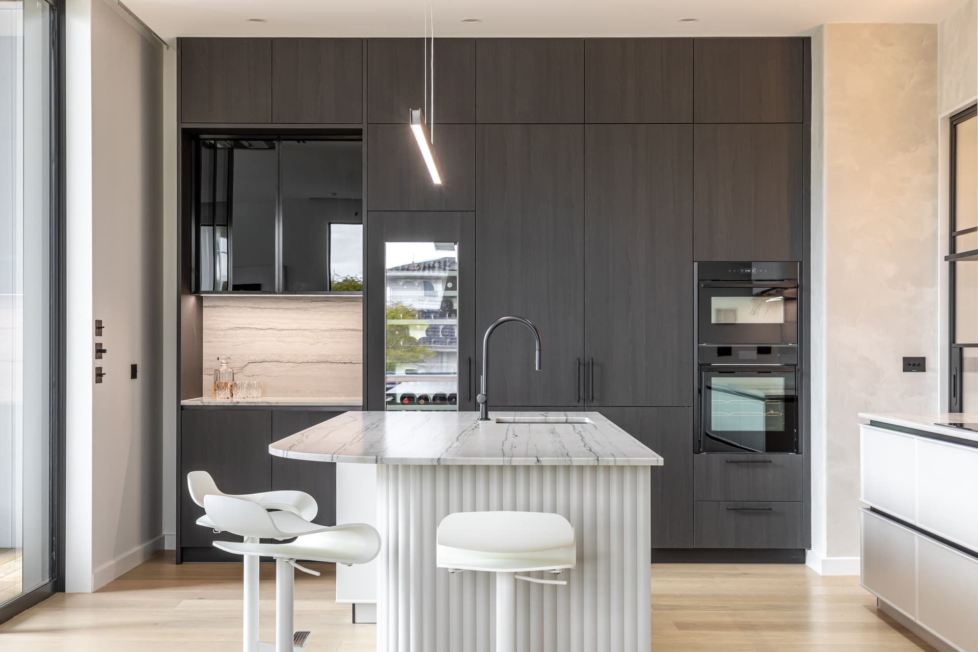 Front-on view of the dark tall cabinet wall, bar niche, wine fridge and ovens behind the island in a Westmere home | Kitchens New Zealand