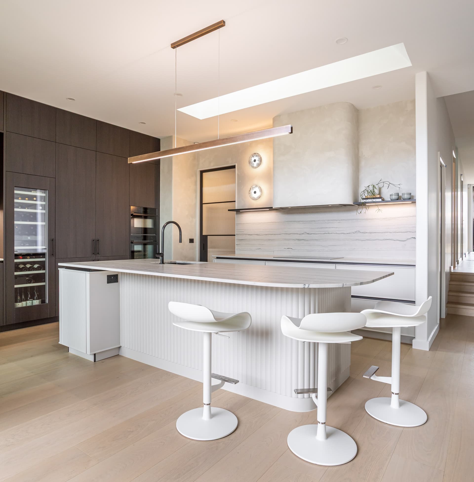 Close view of three white stools at the curved fluted island under a bronze-look linear pendant in a Westmere home | Kitchens New Zealand