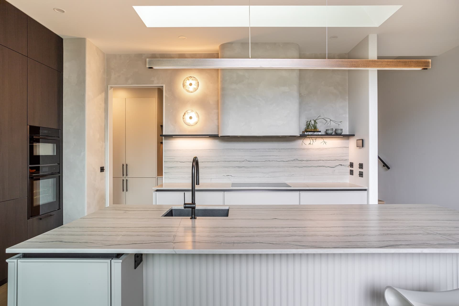 Island sink and black tap facing a curved plaster-look rangehood under a long linear pendant in a Westmere home | Kitchens New Zealand
