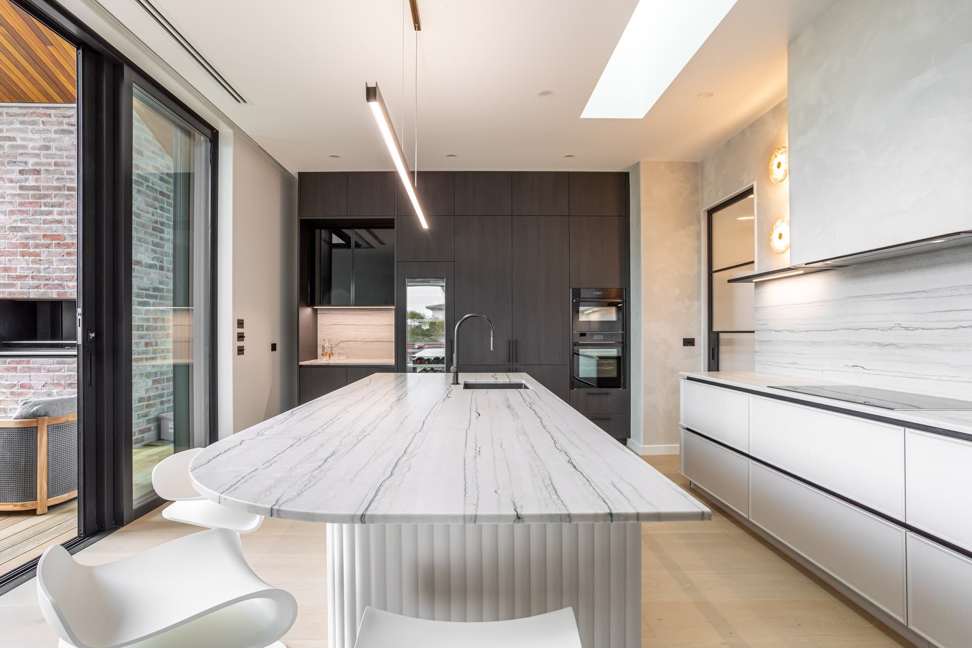 View down the stone-look island to dark tall cabinets, a slider open to a brick-walled deck, in a Westmere home | Kitchens New Zealand