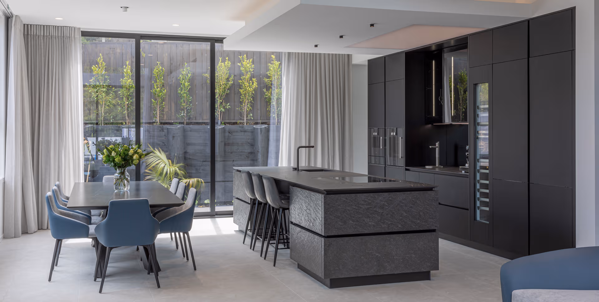 Monochrome kitchen renovation combining matte black finishes, tactile stone and seamless dining integration.