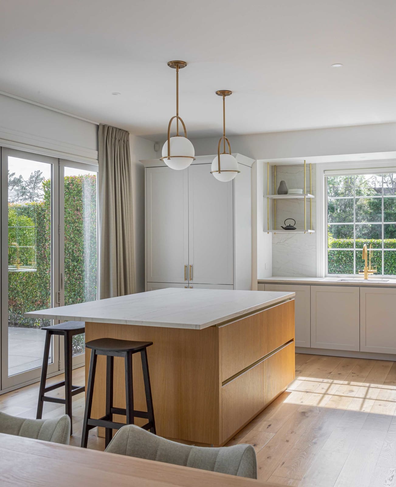 Sunlight across a fluted timber island under brass globe pendants, with glazed doors to the garden