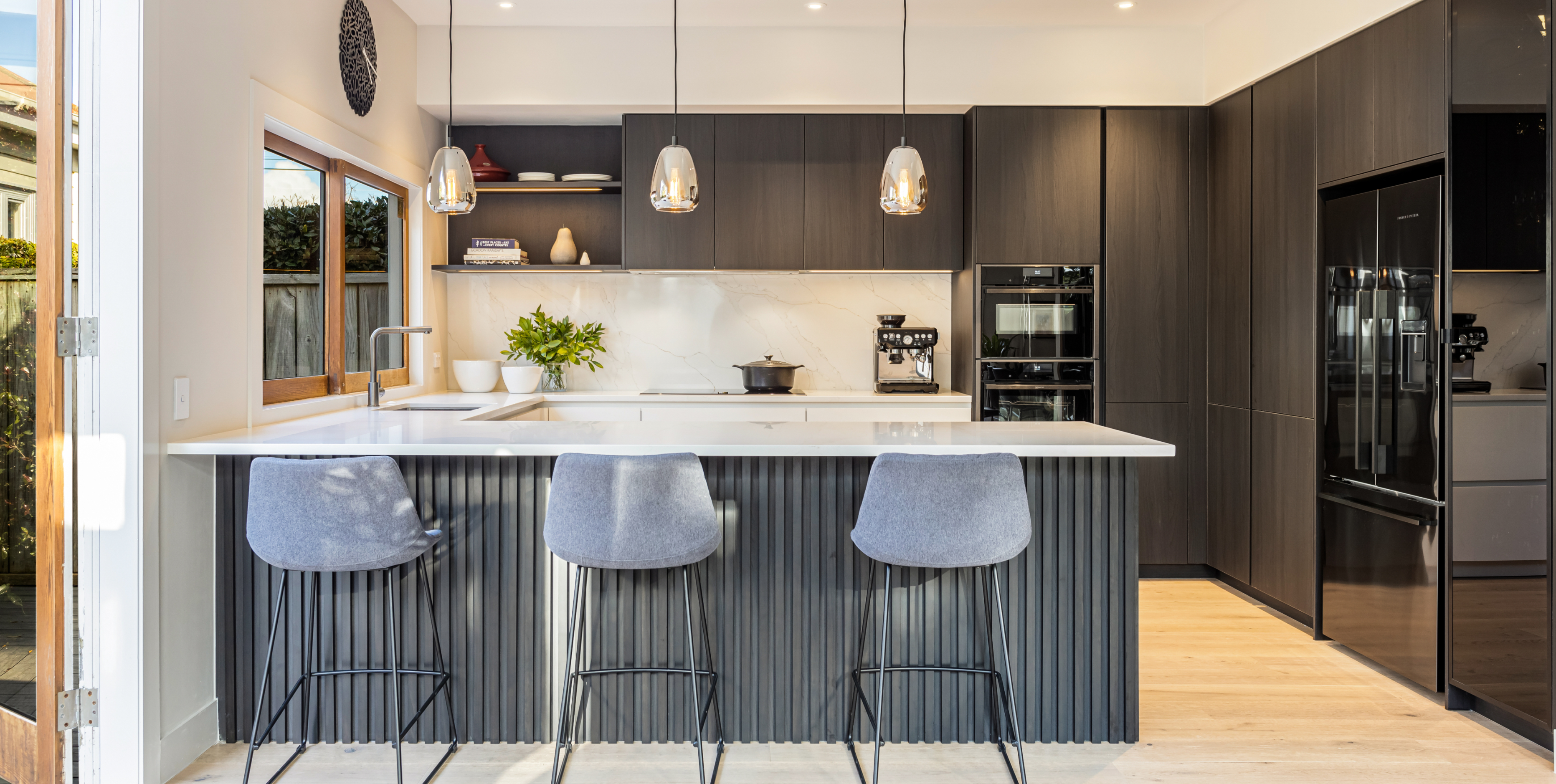 A fluted island anchors the room; bright worktops, dark cabinetry and pendants highlight clean lines and natural light.